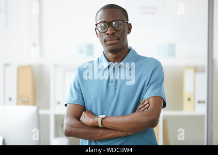 Ritratto di African fiducioso lavoratore di ufficio in occhiali e in maglietta blu in piedi con le braccia incrociate e guardando la telecamera in ufficio Foto Stock