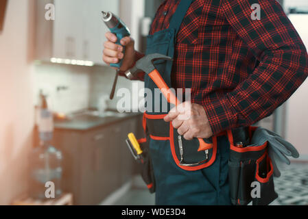 Ritagliato colpo di Uomo in camicia a scacchi indossando tool kit in piedi in cucina, tenendo in mano un martello ed una punta a forare nelle sue mani. Inquadratura orizzontale Foto Stock