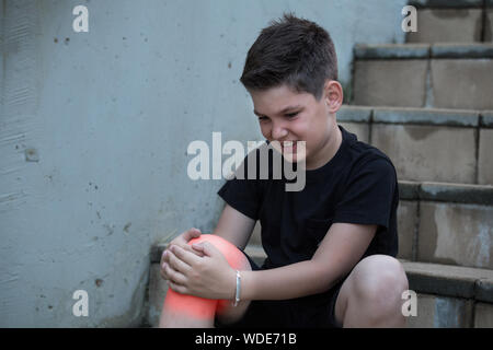 Ragazzo nel dolore guardando la sua gamba ferita seduta sulle scale. Il dolore al ginocchio Foto Stock