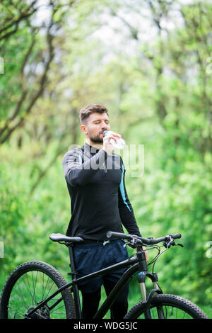 Stanco biker in appoggio e bere da una bottiglia d'acqua. Foto Stock