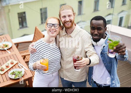 Ritratto di felice giovani amici con cocktail che abbraccia ogni altra e sorridente alla telecamera mentre hanno un pranzo in outdoor cafe Foto Stock