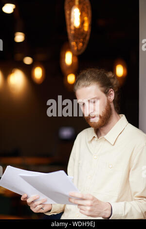 Grave rosso giovane uomo dai capelli tenendo i documenti nelle sue mani ed esaminarli permanente, mentre presso il ristorante Foto Stock