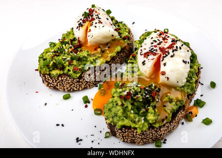 Le uova in camicia con avocado guacomole sul pane marrone con semi di sesamo. Una sana prima colazione su sfondo bianco Foto Stock