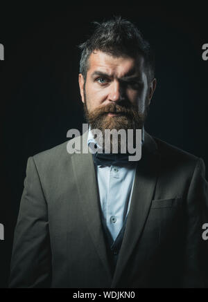 Hipster con aspetto alla moda guarda avanti. Uomo con barba e baffi o barbiere su sfondo scuro. Ragazzo con faccia grave nel lusso abito classico Foto Stock