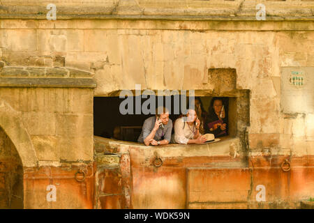 Bagno, Inghilterra - Luglio 2019: visitatori l'ascolto di audio guide peer attraverso una parete del bagno romano nel centro della città di Bath Foto Stock