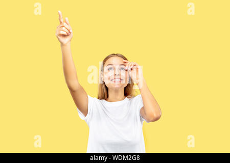 Giovani caucasici donna di mezza lunghezza verticale sul giallo di sfondo per studio. Bellissimo modello femminile in camicia bianca. Concetto di emozioni umane, l'espressione del viso. Dispositivo di puntamento su e sorridente, si chiedeva. Foto Stock