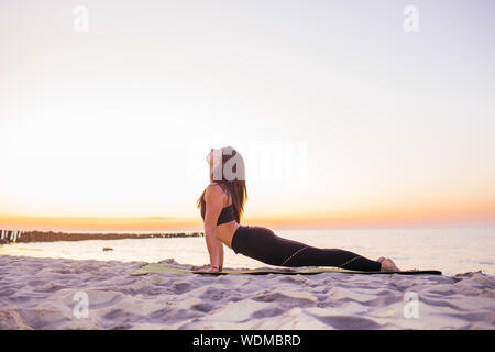 Giovani slim yogi donna facendo Urdhva Mukha Svanasana o rivolto verso l'alto cane pongono contro il tramonto sulla spiaggia. Healhy concetto di stile di vita Foto Stock