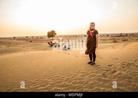 Una donna su un cammello trek tour nel deserto di Thar di Rajasthan orientale a guardare il tramonto con i suoi cammelli e driver / guide. Rajasthan, India. Foto Stock