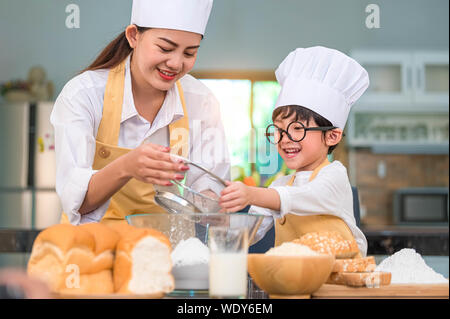 Grazioso piccolo ragazzo asiatico e madre bella pasta setacciare la farina con il setaccio dello spolverizzatore scolapasta nella cucina di casa sul tavolo per preparare al forno di cottura e ca Foto Stock