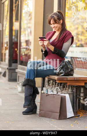 Felice, sorridente, giovane donna ispanica con borse per lo shopping e il telefono cellulare. Guarda come se i messaggi di testo, il controllo della posta elettronica o utilizzando un'applicazione. Foto Stock
