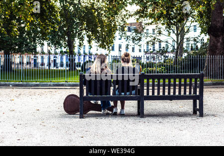 Vista posteriore di un paio di giovani donne seduto su una panca in legno a Fitzroy Square Garden, Fitzrovia, Londra, Inghilterra, Regno Unito. Foto Stock
