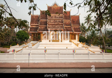 Il 4 aprile, 2019 Luang Prabang, Laos - Luang Prabang Royal Palace Museum e Hor Prabang temple hall visto throuhg rami di alberi con vuoto street in mornin Foto Stock