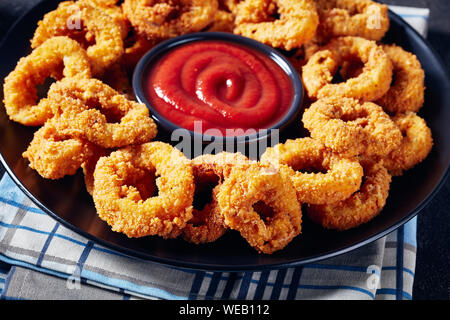 Croccanti anelli di calamari e deep-panate anelli di totano servito con salsa di pomodoro su una piastra nera su un tovagliolo, vista orizzontale dal di sopra, close-up Foto Stock