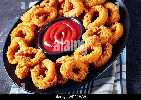 Close-up di croccanti anelli di calamari e deep-panate anelli di totano servito con salsa di pomodoro su una piastra nera su un tovagliolo, orizzontale vista da sopra Foto Stock