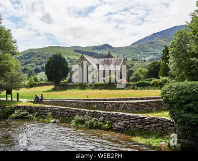 St Marys chiesa in Beddgelert in Snowdonia su una luminosa giornata d'estate con due persone sedute su un muro di pietra accanto al fiume Glaslyn, Wales, Regno Unito Foto Stock