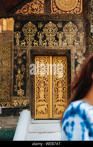 APR 5 Luang Prabang, Laos - Golden Buddismo arte murale di legno intagliato gate con la donna in primo piano a sala principale di Wat Xieng Thong Foto Stock