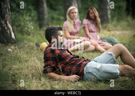 Uomo Barbuto giacente in erba sul prato boschivo. Ragazze seduto accanto al fuoco. Amici in campeggio in tarda estate. Relax nella foresta. Foto Stock