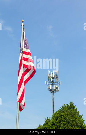 Una bandiera americana sulla bandiera pole affiancato ad un telefono cellulare la torre e il picco di un albero sempreverde. Cielo blu sullo sfondo. Copia dello spazio. Foto Stock