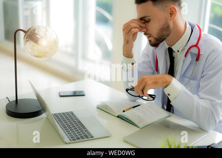 Sottolineato medico seduto alla sua scrivania nel suo ufficio mentre lui overworke presso l'ospedale. Medicina, sanità e concetto di persone. Foto Stock