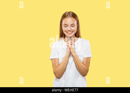 Giovani caucasici donna di mezza lunghezza verticale sul giallo di sfondo per studio. Bellissimo modello femminile in camicia bianca. Concetto di emozioni umane, espressione facciale, vendite. Pregando con gli occhi chiusi, sorridente. Foto Stock