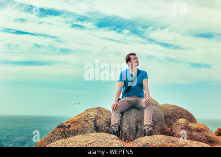 Uomo seduto su di una roccia al mare guardando in alto Foto Stock
