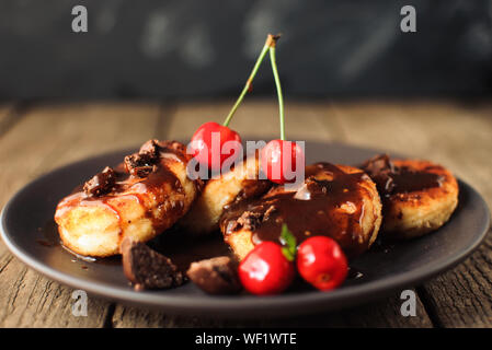 Colazione Gourmet - frittelle di cagliata, cheesecake, cagliata frittelle con ciliegie e cioccolato in una piastra di marrone. Salubre dessert su una tavola di legno in un r Foto Stock