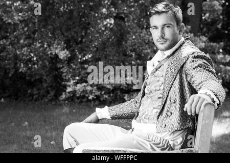 Ritratto di uomo bello vestito in costume d'epoca seduta sul banco in giardino Foto Stock