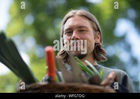 Ritratto di un uomo in piedi in un giardino che porta un cesto pieno di attrezzature da giardinaggio, Germania Foto Stock