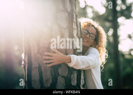 Bella donna con gli occhiali che abbraccia un grande albero con amore e con affetto - Salvare il concetto di foresta e stile di vita - per adulti in montagna o in un parco Foto Stock