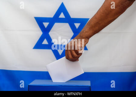 Uomo israeliano di mettere un voto in un urna elettorale il giorno delle elezioni. Stretta di mano con voti bianco carta sulla bandiera di Israele lo sfondo. Mockup, spazio per il testo. Foto Stock