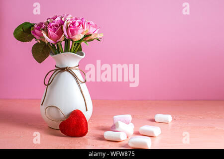 Di piccole dimensioni e di colore bianco e rosa marshmallows sono sparsi su un pallido sfondo rosa accanto a un vaso di rose. Posto per il testo. Foto Stock