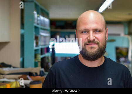 Ritratto di un muso da parte calva con la barba, calzolaio cercando fotocamera presso la sua officina dolce sorriso. Foto Stock