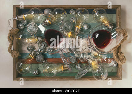 Due bicchieri di vino rosso e retrò ghirlanda di Natale con le lampade accese nel vassoio in legno su marmo bianco dello sfondo. Laici piana, spazio Foto Stock