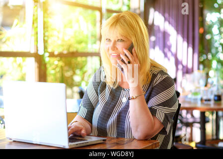 Ritratto di felice occupato vendite più dimensione donna seduta al cafe e utilizzando il suo smartphone. Donna di affari di lavoro on-line. Attraente imprenditrice maturo lavorando sul computer portatile in area di lavoro. Foto Stock