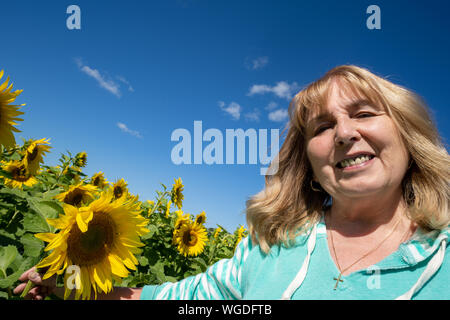 Senior donna pone in un campo di girasoli, sorridente contro un luminoso cielo blu Foto Stock