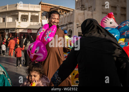 Doha, Qatar - 17 DIC 2016: un uomo street vendor è guardando la telecamera e la vendita di un palloncino colorato per una ragazza e una donna che indossa un tradizionale ab Foto Stock