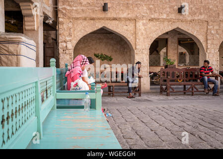 Doha, Qatar - 3 DIC 2016: due donne dalle Filippine e due indiani gli uomini sono seduti sulla città sul turchese panchine nei Souq Wakif, Doha Foto Stock