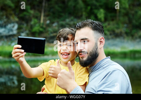 Family photo shoot in natura. Padre e figlio prendere un giunto selfie, essi stessi fotografia sulle rive di un fiume selvaggio, una telecamera in un telefono cellulare. Foto Stock