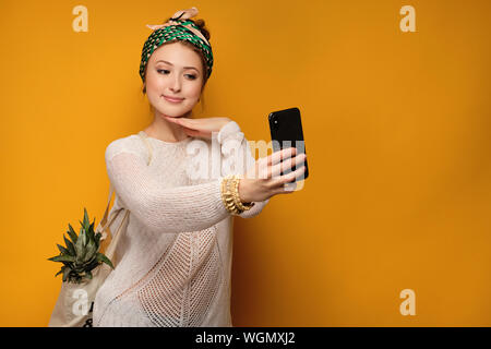 Ragazza in un maglione leggero e annodate una testa colorata sciarpa con un ananas in una borsa e prende un selfie. Foto Stock