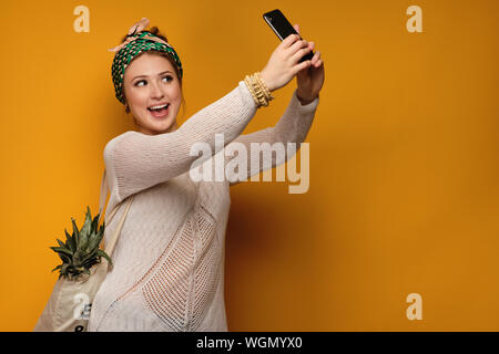 Ragazza in un maglione leggero e annodate una testa colorata sciarpa con un ananas in un sacchetto e sorridente felicemente rende selfies. Foto Stock