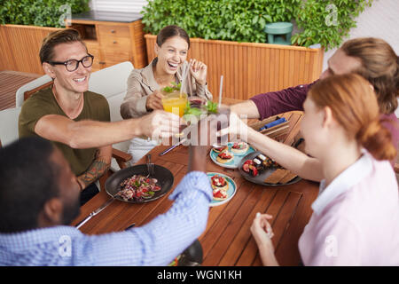 Angolo di alta vista sul gruppo di persone contemporanee tostatura e bicchieri tintinnanti durante la celebrazione della vacanza a cena in cafe, spazio di copia