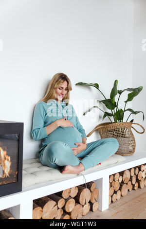 Ragazza incinta in abbigliamento sportivo verde avvolge il bambino non ancora nato e le pratiche yoga, in interni accoglienti Foto Stock