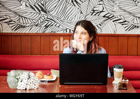 Fantasticando giovane donna seduta al tavolo del bar aperto con laptop, caffè, pasticceria e bouquet di fiori e guardando attraverso la finestra Foto Stock