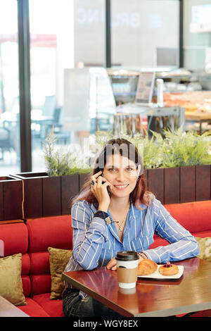 Ritratto di pretty sorridente imprenditrice seduti al tavolo del bar, avente tazza di caffè con soffi e parlando al telefono Foto Stock