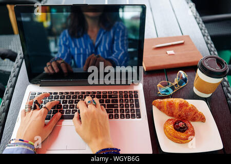 Prospettiva in prima persona di sesso femminile di imprenditore di mangiare la prima colazione di caffè e pasticceria e di lavoro sul computer portatile Foto Stock