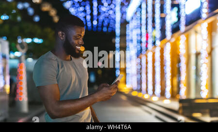 Sorridente uomo nero tenendo lo smartphone più recenti, a piedi dalla città di notte Foto Stock