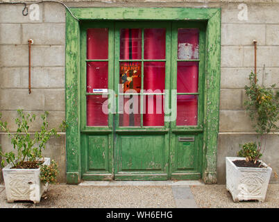 Vecchio edificio con weathered green in pannelli di vetro della porta di legno, tende rosa, su entrambi i lati i vasi da fiori in plastica con piante avvizzite, un poster di calcio Foto Stock