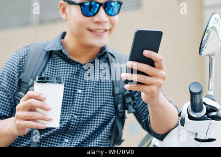Immagine ritagliata positiva di giovane uomo asiatico la lettura di messaggi di testo sullo smartphone quando si è seduti all'aperto Foto Stock