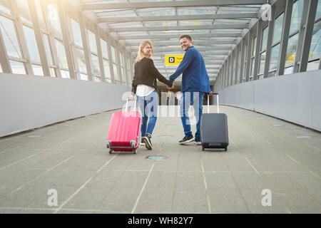 Foto dal retro di un uomo e di una donna che guarda la fotocamera, a piedi con le valigie sul passaggio all'aeroporto durante il giorno Foto Stock