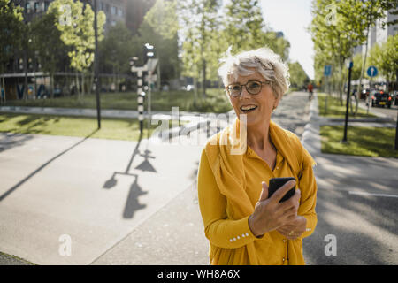 Ritratto di felice donna matura con lo smartphone che indossa abiti di colore giallo all'aperto Foto Stock